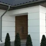 Nowoczesny dom z białymi panelami, drewnianymi drzwiami i rynną. Wokół rosną tuje, tworząc miły bonio.
