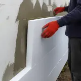 Ręce w czerwonych rękawicach przytwierdzają płyty styropianowe do ściany, rozpoczynając ocieplanie elewacji.