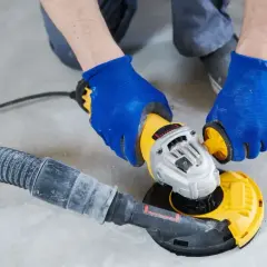 Ręce w niebieskich rękawiczkach prowadzą szlifierkę z odkurzaczem, wykonując szlifowanie betonu.