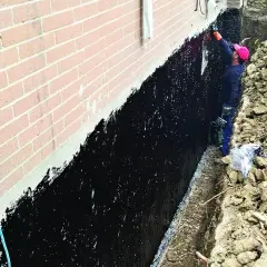 Pracownik zabezpiecza ławy fundamentowe czarną masą izolacyjną.