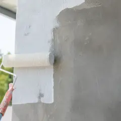 Wałek malarski pokrywa białą farbą szary beton. Prace wykończeniowe na zewnątrz budynku.