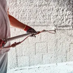 Pracownik w rękawicach nakłada obrzutkę cementową na ścianę za pomocą agregatu.