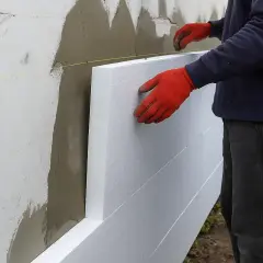 Ręce w czerwonych rękawicach przytwierdzają płyty styropianowe do ściany, rozpoczynając ocieplanie elewacji.