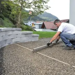 Mężczyzna wyrównuje żwir, tworząc podjazd z odpowiednim odprowadzeniem wody. Obok wznosi się mur oporowy z bloczków.