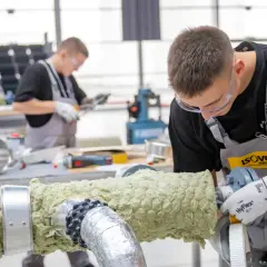 Młody mężczyzna w okularach ochronnych i fartuchu pracuje nad izolacją rury. Dbałość o detale i precyzja zapewniają dobrą adhezję materiału izolacyjnego.