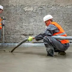 Dwóch budowlańców w kamizelkach odblaskowych wylewa beton. Jeden kieruje wąż z masą, drugi kontroluje poziom.
