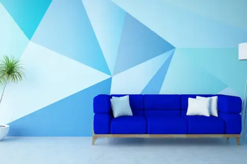 Niebieska sofa i roślina w donicy na tle ściany z geometrycznym wzorem, tworzącym efekt malarski.