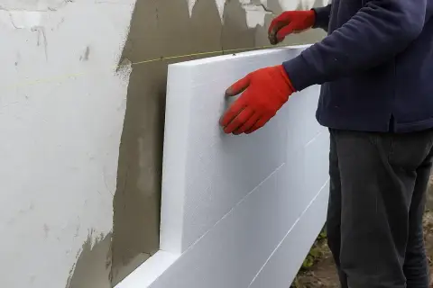 Ręce w czerwonych rękawicach przytwierdzają płyty styropianowe do ściany, rozpoczynając ocieplanie elewacji.