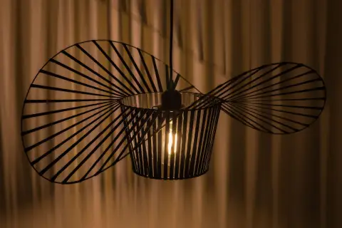Nowoczesna lampa wisząca o nietypowym kształcie, przypominającym wachlarz lub wachlarz, tworzy ciekawe cienie na zasłonie.