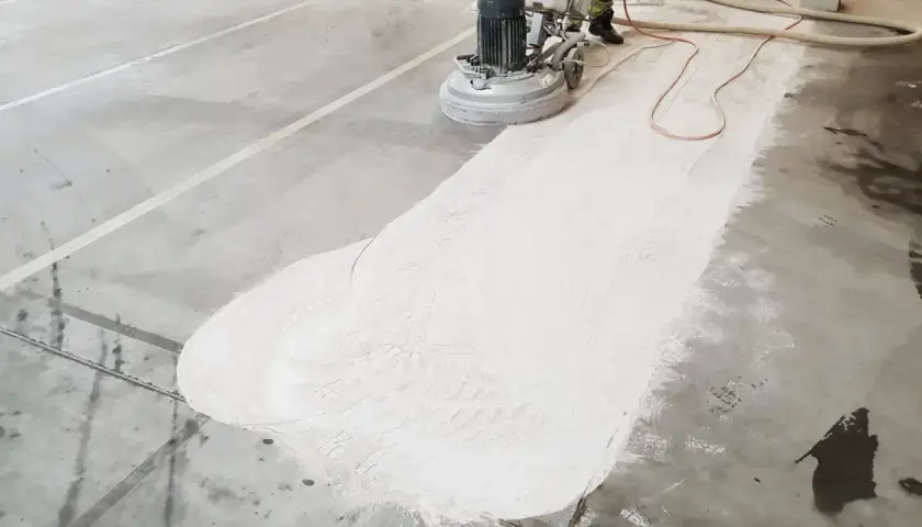 Maszyna szlifuje betonową posadzkę, tworząc biały pył. Trwa naprawa posadzki betonowej, przygotowanie do dalszych prac.