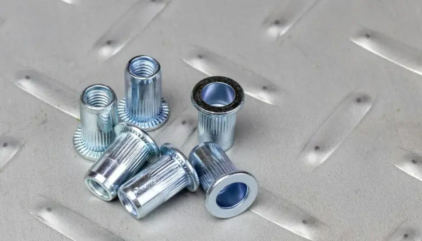 Kilka metalowych nitonakrętek leży na chropowatej, metalowej powierzchni.