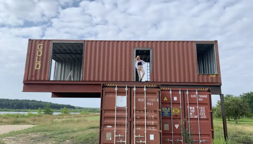 Kobieta stoi w drzwiach domu z kontenera morskiego, który jest częścią nietypowej budowli.