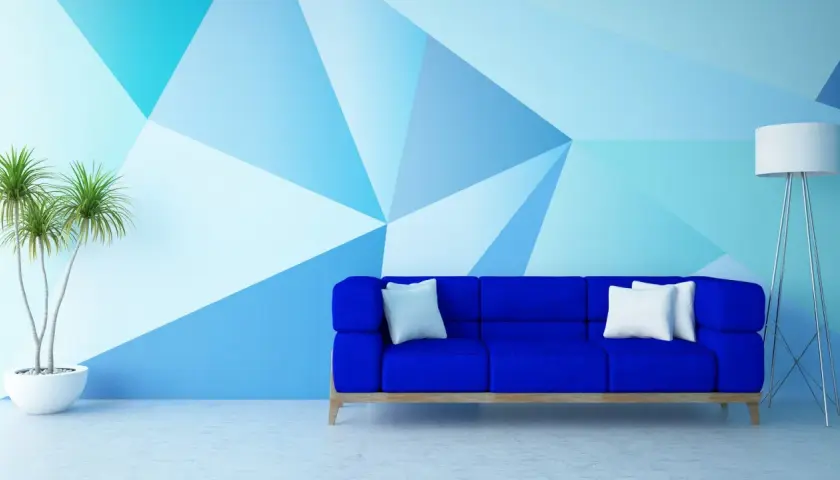 Niebieska sofa i roślina w donicy na tle ściany z geometrycznym wzorem, tworzącym efekt malarski.