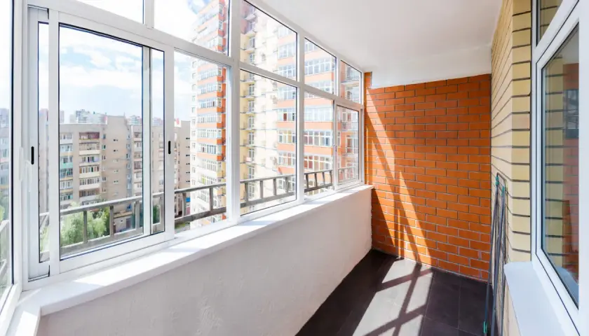 Szklana loggia z widokiem na blokowisko. Jasny balkon z białymi oknami i ceglaną ścianą.