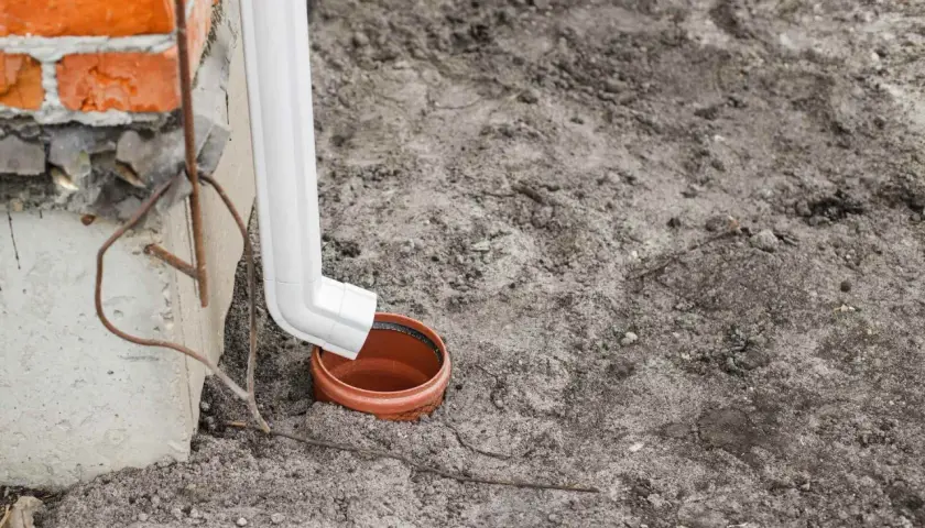Biała rura odprowadza wodę z rynny do brązowej studzienki. Ceglany mur i betonowa podstawa budynku.