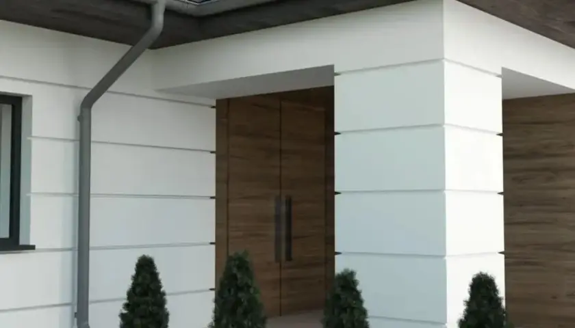 Nowoczesny dom z białymi panelami, drewnianymi drzwiami i rynną. Wokół rosną tuje, tworząc miły bonio.