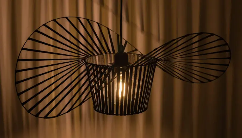 Nowoczesna lampa wisząca o nietypowym kształcie, przypominającym wachlarz lub wachlarz, tworzy ciekawe cienie na zasłonie.