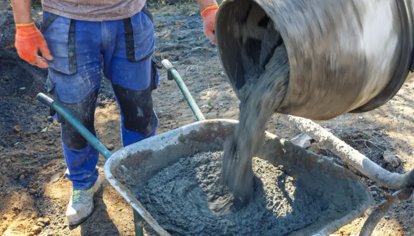 Robotnik w pomarańczowych rękawicach wylewa świeży beton z betoniarki do taczki. Różne rodzaje betonu wymagają różnych metod mieszania i transportu.