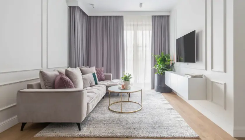 Elegancki salon w stylu klasycznym. Jasna sofa, okrągły stolik kawowy, dywan i zasłony tworzą przytulną atmosferę.