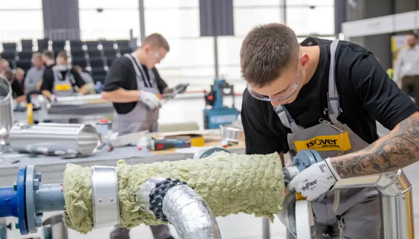 Młody mężczyzna w okularach ochronnych i fartuchu pracuje nad izolacją rury. Dbałość o detale i precyzja zapewniają dobrą adhezję materiału izolacyjnego.