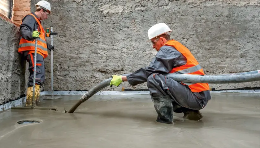 Dwóch budowlańców w kamizelkach odblaskowych wylewa beton. Jeden kieruje wąż z masą, drugi kontroluje poziom.
