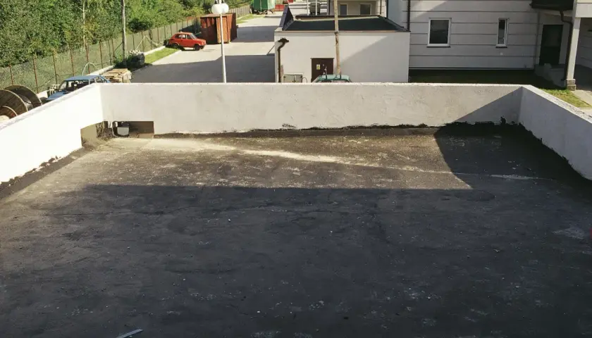 Widok na szary, płaski dach z białymi ściankami. W tle budynki, drzewa i czerwony samochód.