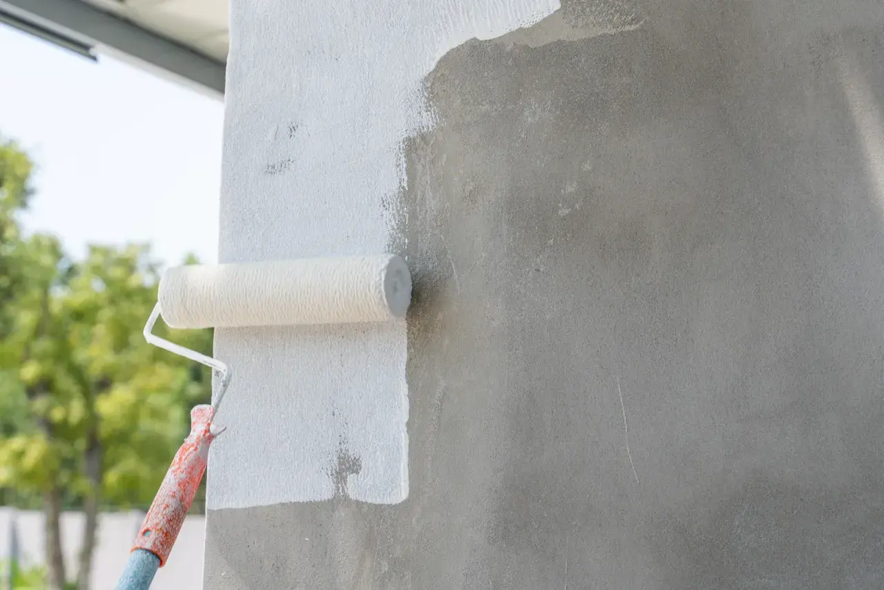 Wałek malarski pokrywa białą farbą szary beton. Prace wykończeniowe na zewnątrz budynku.