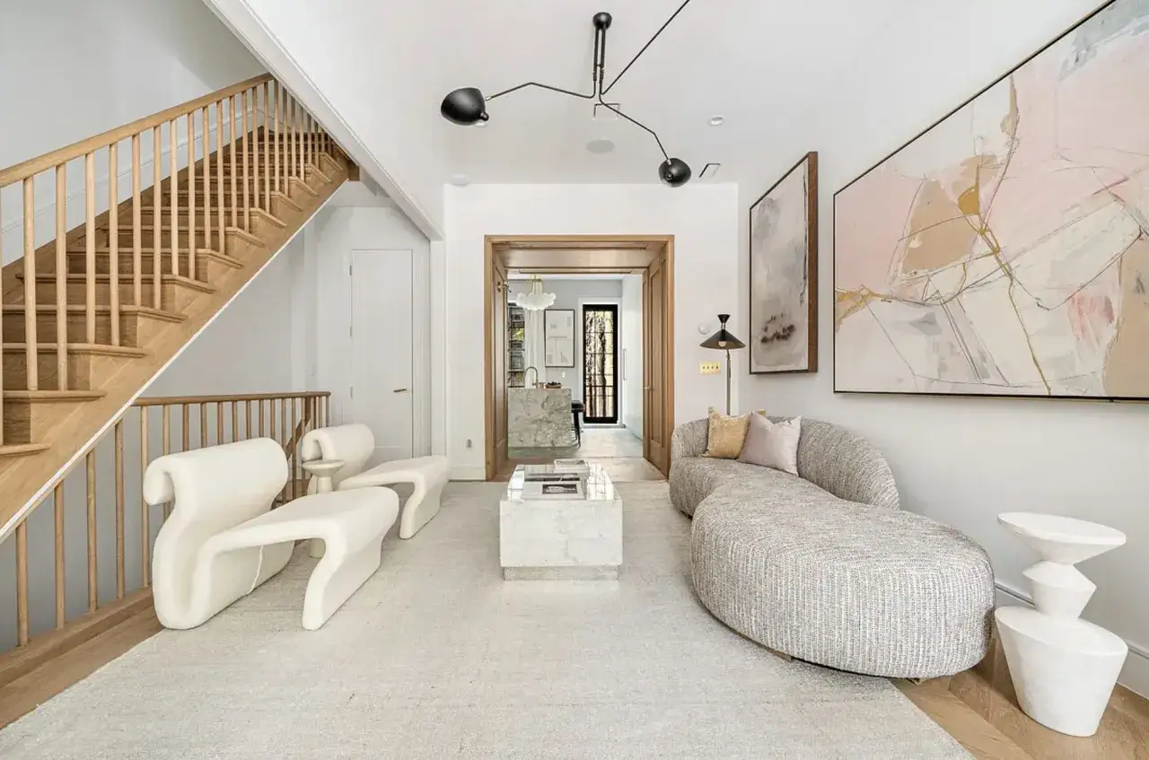 Nowoczesny salon w stylu minimalistycznym. Jasne meble, drewniane schody, abstrakcyjne obrazy i designerska lampa tworzą elegancką przestrzeń.