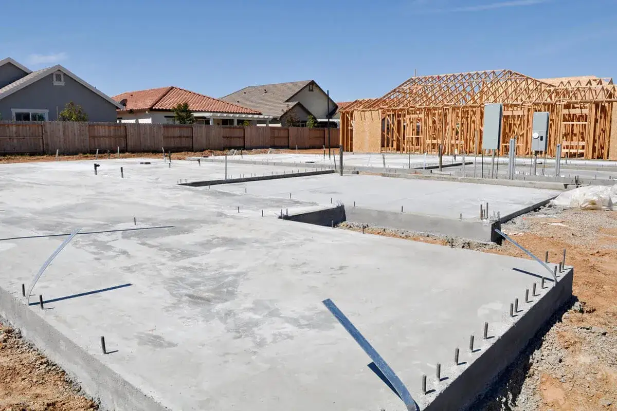 Fundament domu z chudego betonu, z widocznymi elementami konstrukcyjnymi i instalacjami. W tle budowane są drewniane szkielety domów.