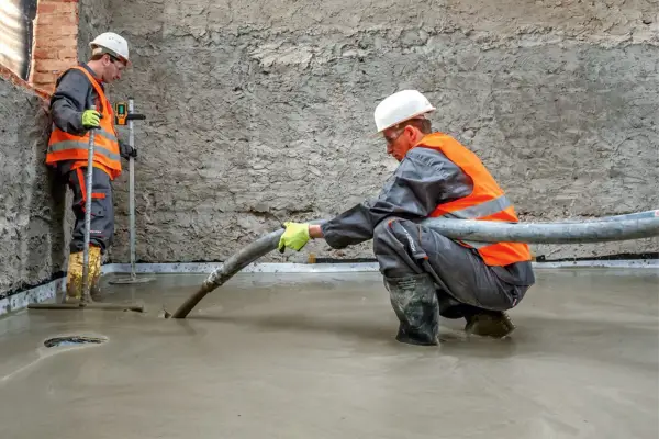 Dwóch budowlańców w kamizelkach odblaskowych wylewa beton. Jeden kieruje wąż z masą, drugi kontroluje poziom.